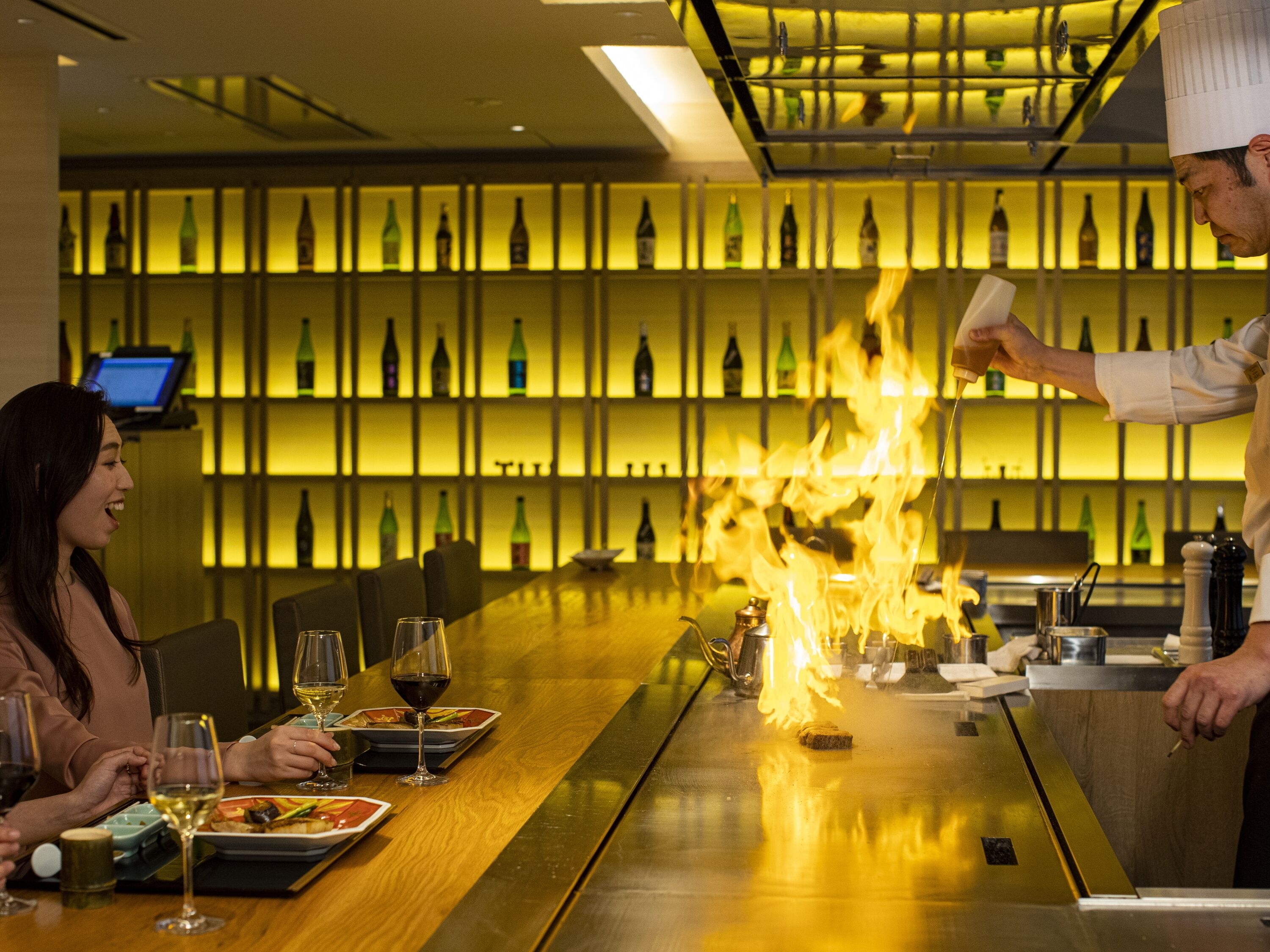 A restaurant with a long table, wine glasses, and a chef performing a flambé.
