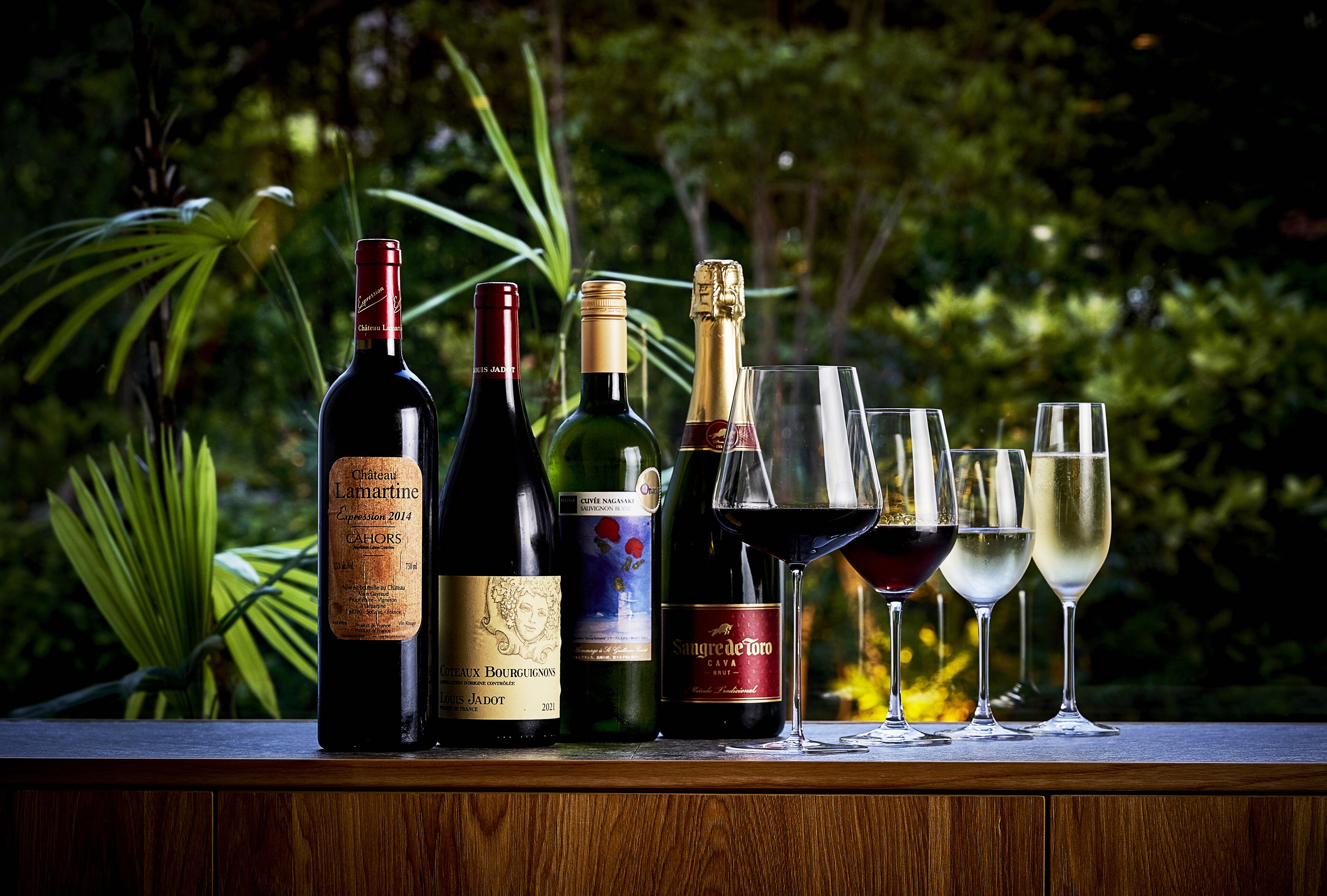 A selection of wine bottles and glasses on a wooden surface with a lush green background.