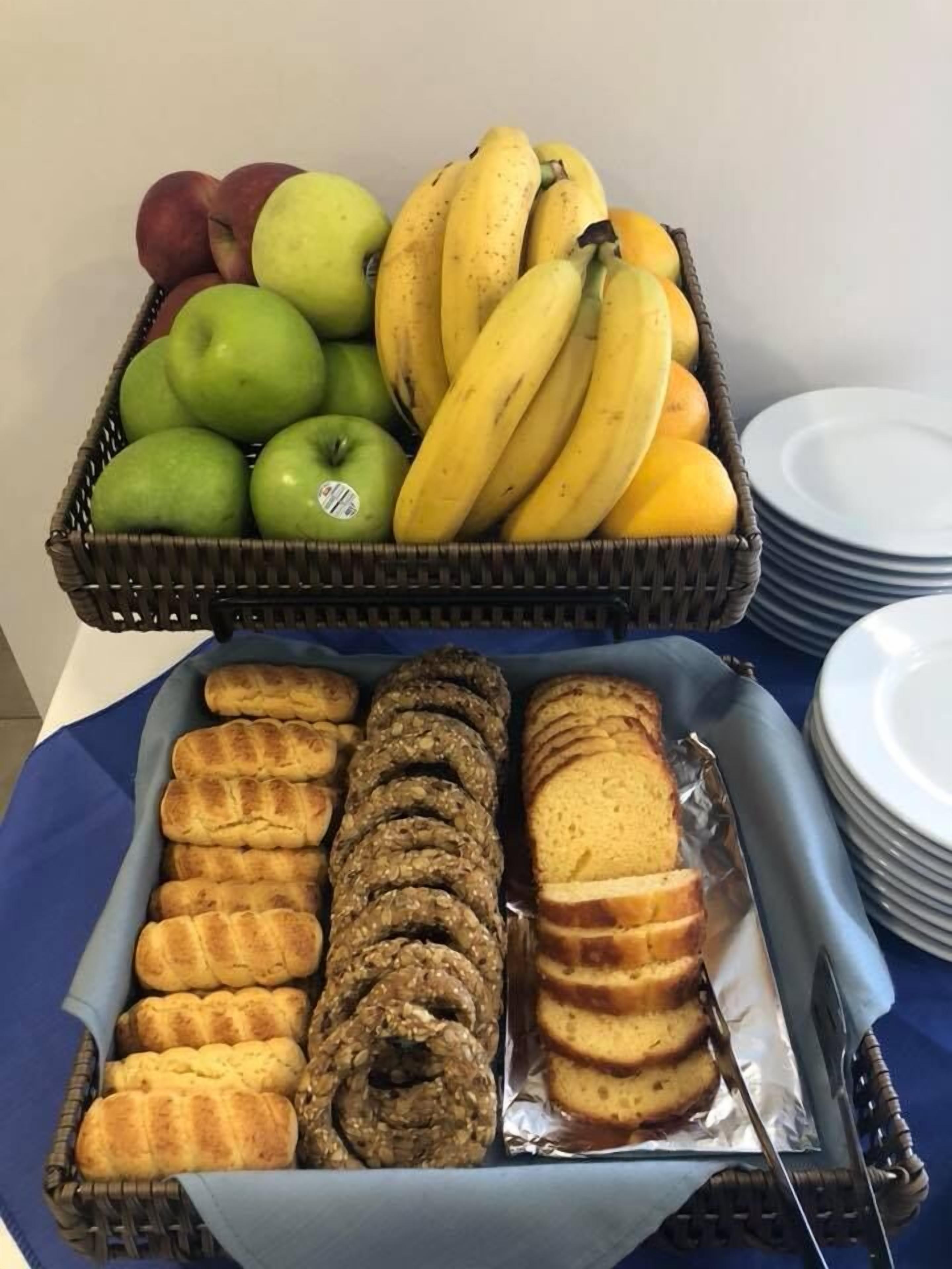 Una cesta de frutas con manzanas y plátanos, y una bandeja con panecillos y pan rebanado.