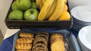 Una cesta de frutas con manzanas y plátanos, y una bandeja con panecillos y pan rebanado.