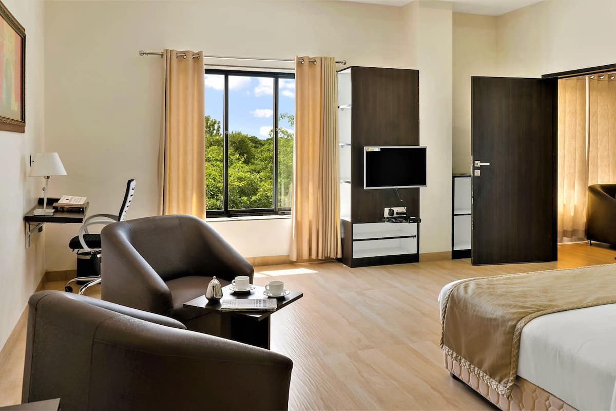 Quarto de hotel com cama, escrivaninha, luminária, poltrona, televisão e janela com cortinas.