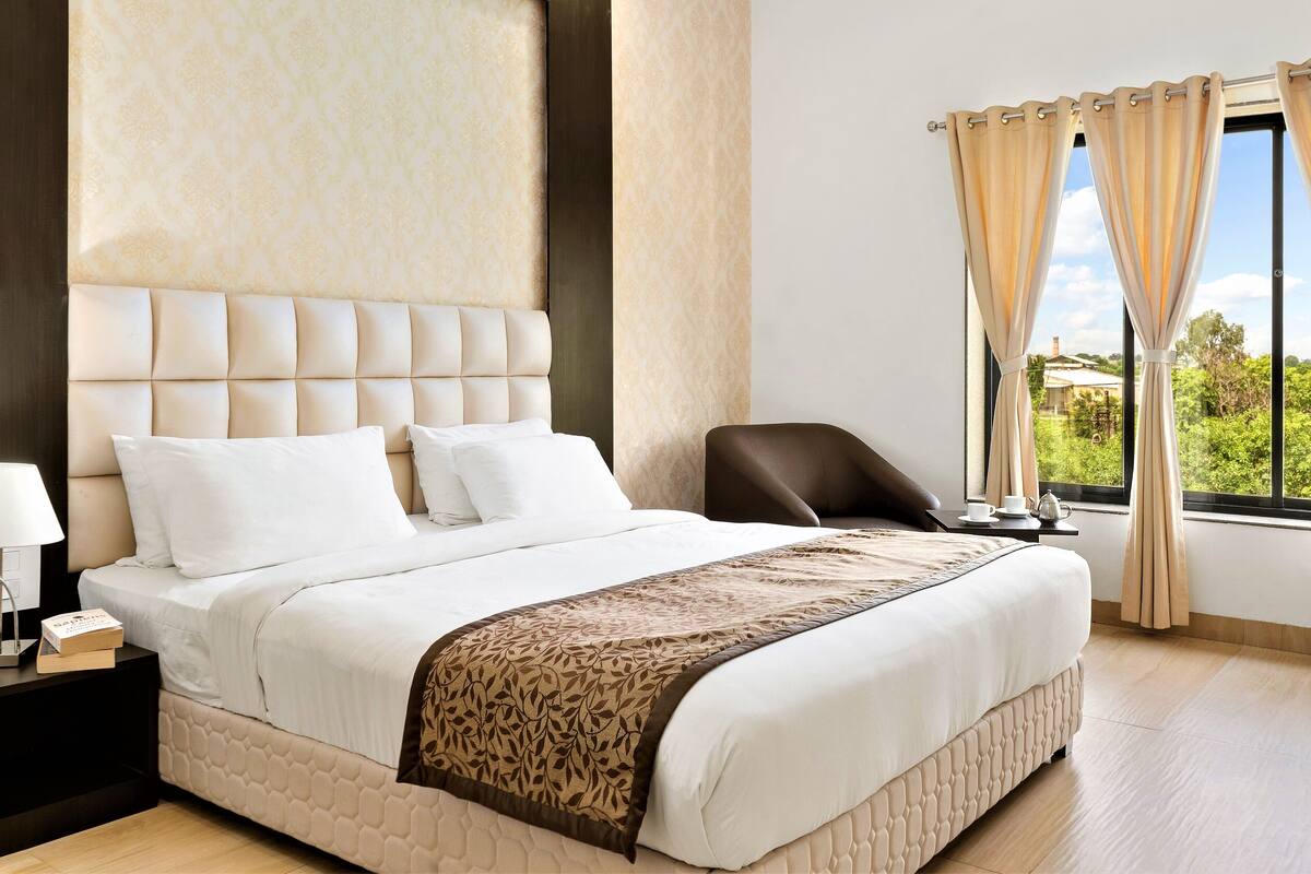 Quarto de hotel com uma cama grande, uma mesa de cabeceira, uma cadeira e uma janela com cortinas.
