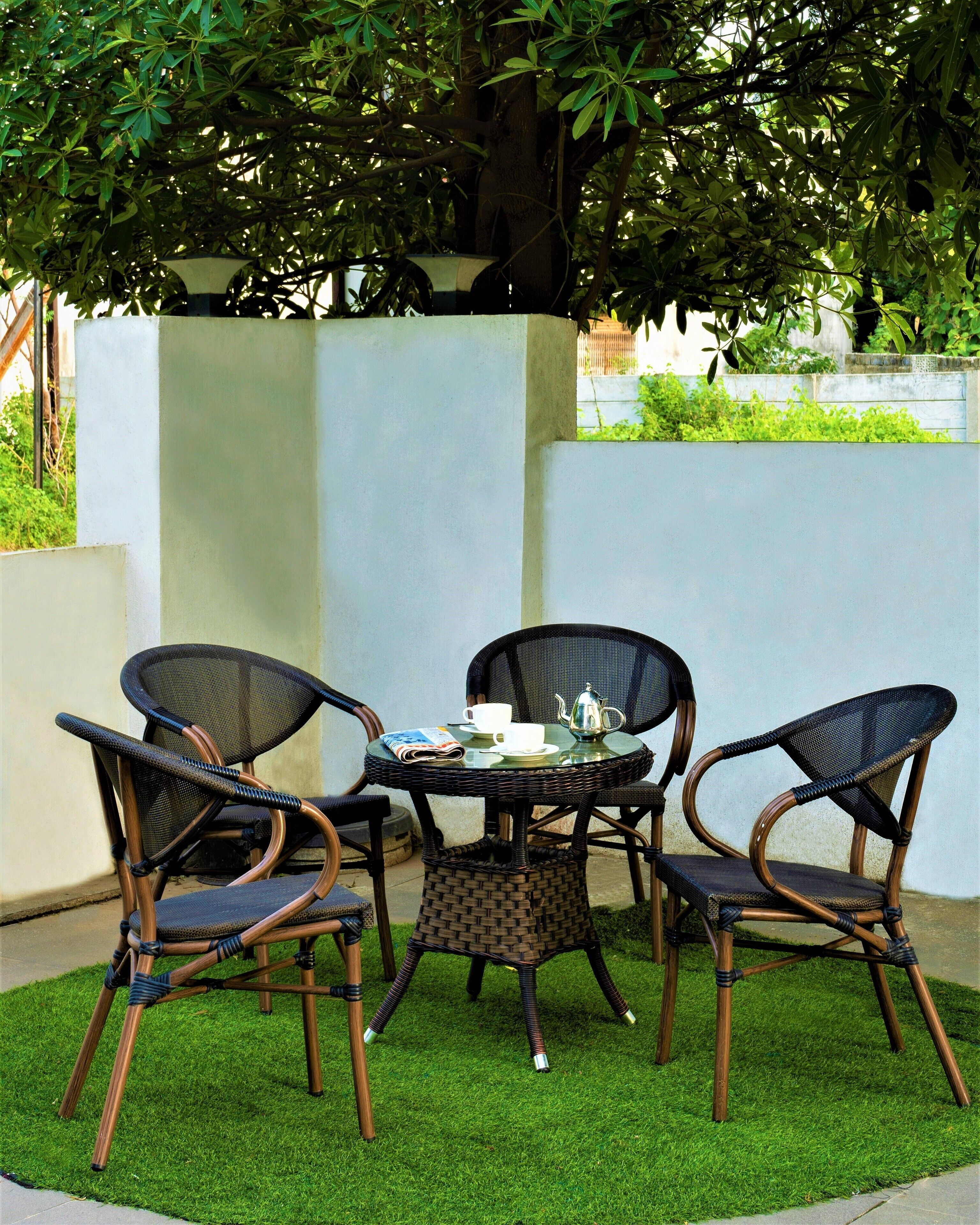 A round table with three wicker chairs, a white cup, and a silver teapot on it.