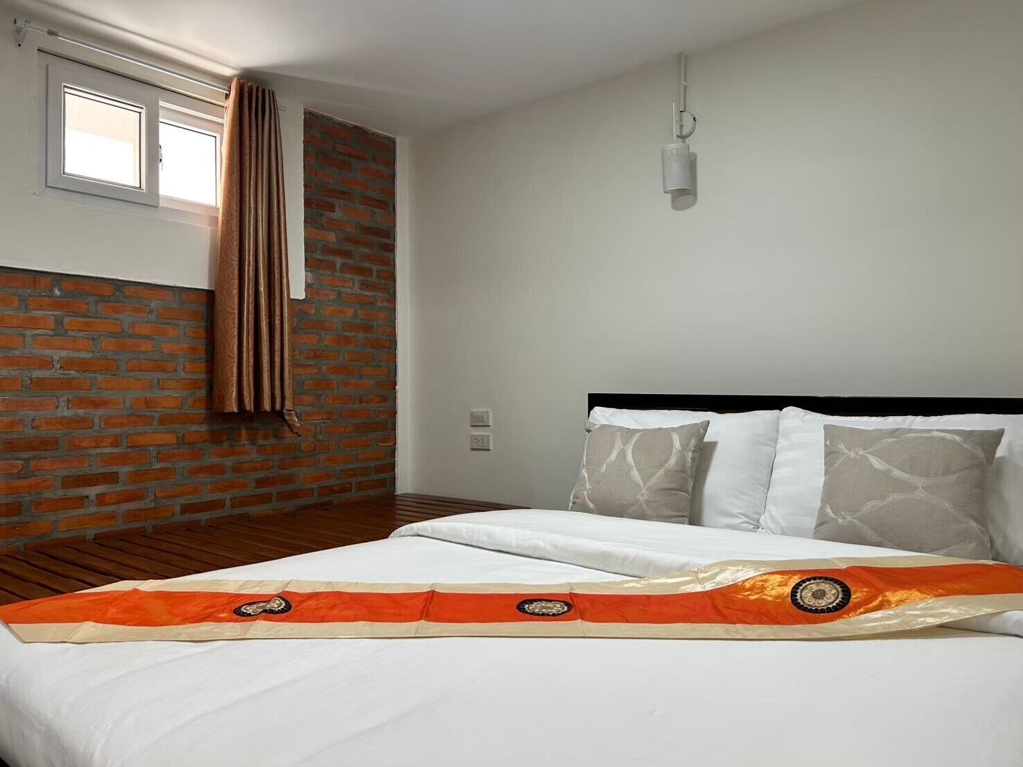 Un dormitorio con pared de ladrillo, ventana con cortinas, cama con ropa blanca y un cabecero decorativo anaranjado y beige.