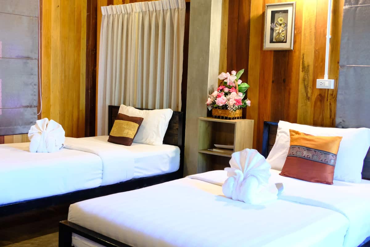 Two single beds with white linens, a wooden headboard, a small bedside table with a vase of flowers, and a curtain.