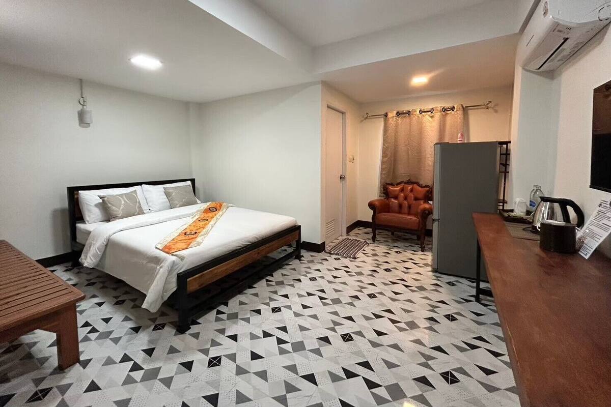 A compact hotel room with a bed, a wooden table, a chair, and a kitchenette area.