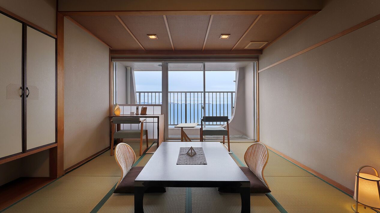 Een kamer met tatami vloer, een lage tafel met kussen, een stoel, een tafeltje met lamp en uitzicht op zee door grote ramen.