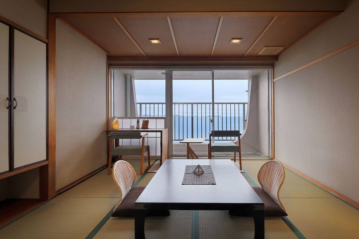 Een kamer met tatami vloer, een lage tafel met kussen, een stoel, een tafeltje met lamp en uitzicht op zee door grote ramen.