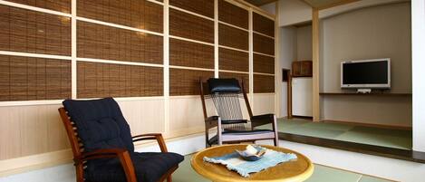 A traditional Japanese room with tatami flooring, a low wooden table, two chairs, and a television on a shelf.