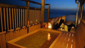 A wooden deck with a hot tub, bamboo fence, and a view of the ocean at dusk.