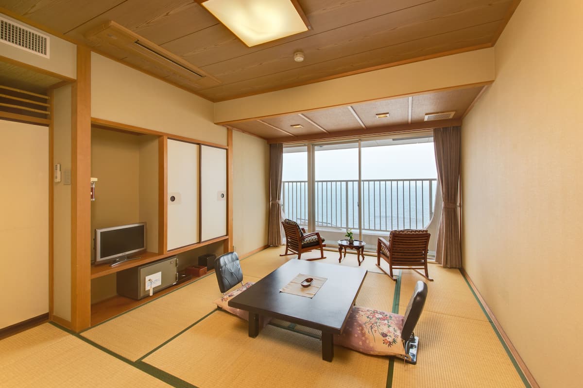 A traditional Japanese room with tatami flooring, a low table, chairs, a television, and a large window with a view of the sea.
