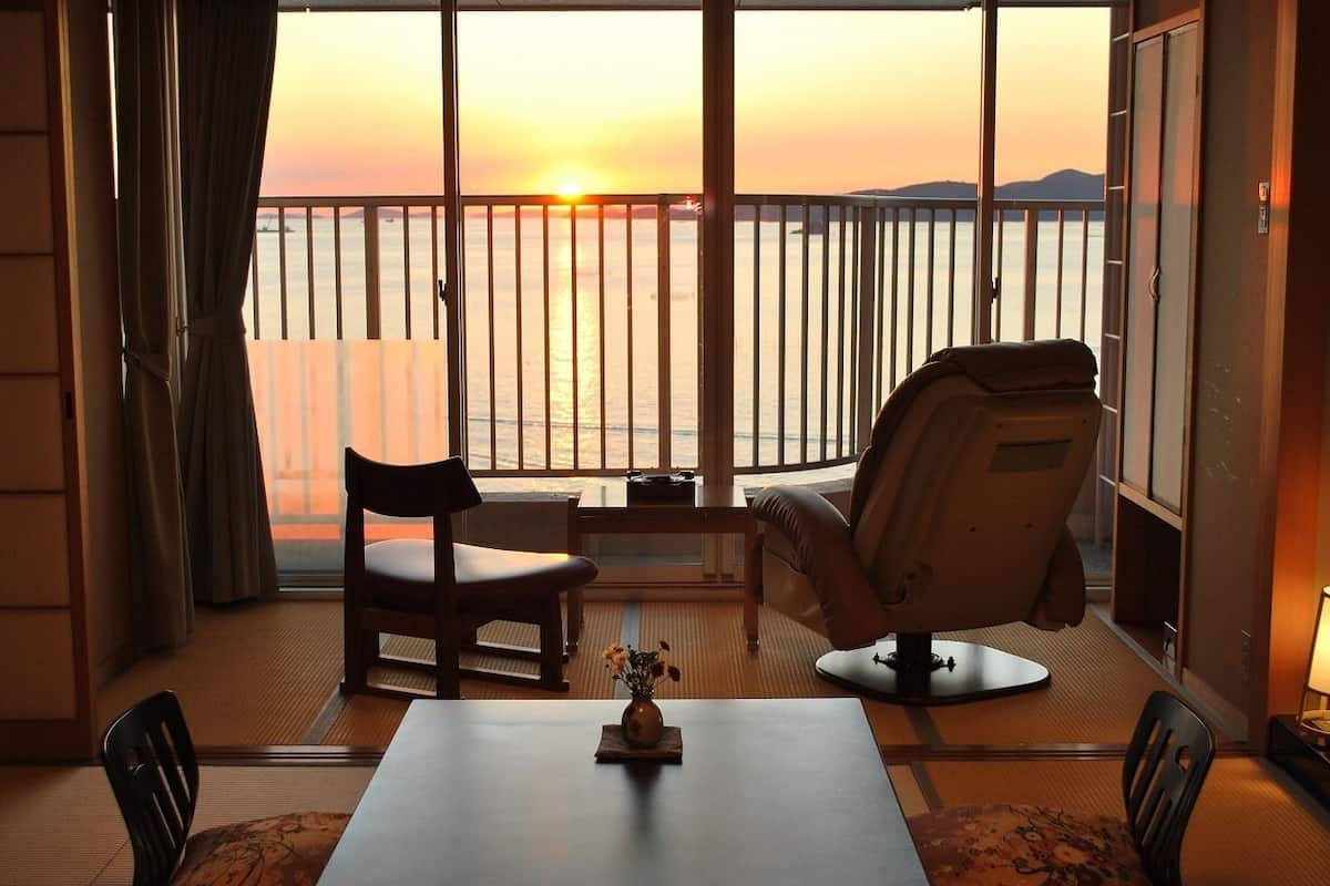 A room with a view of the sunset over the water, a table with a vase, and two chairs.