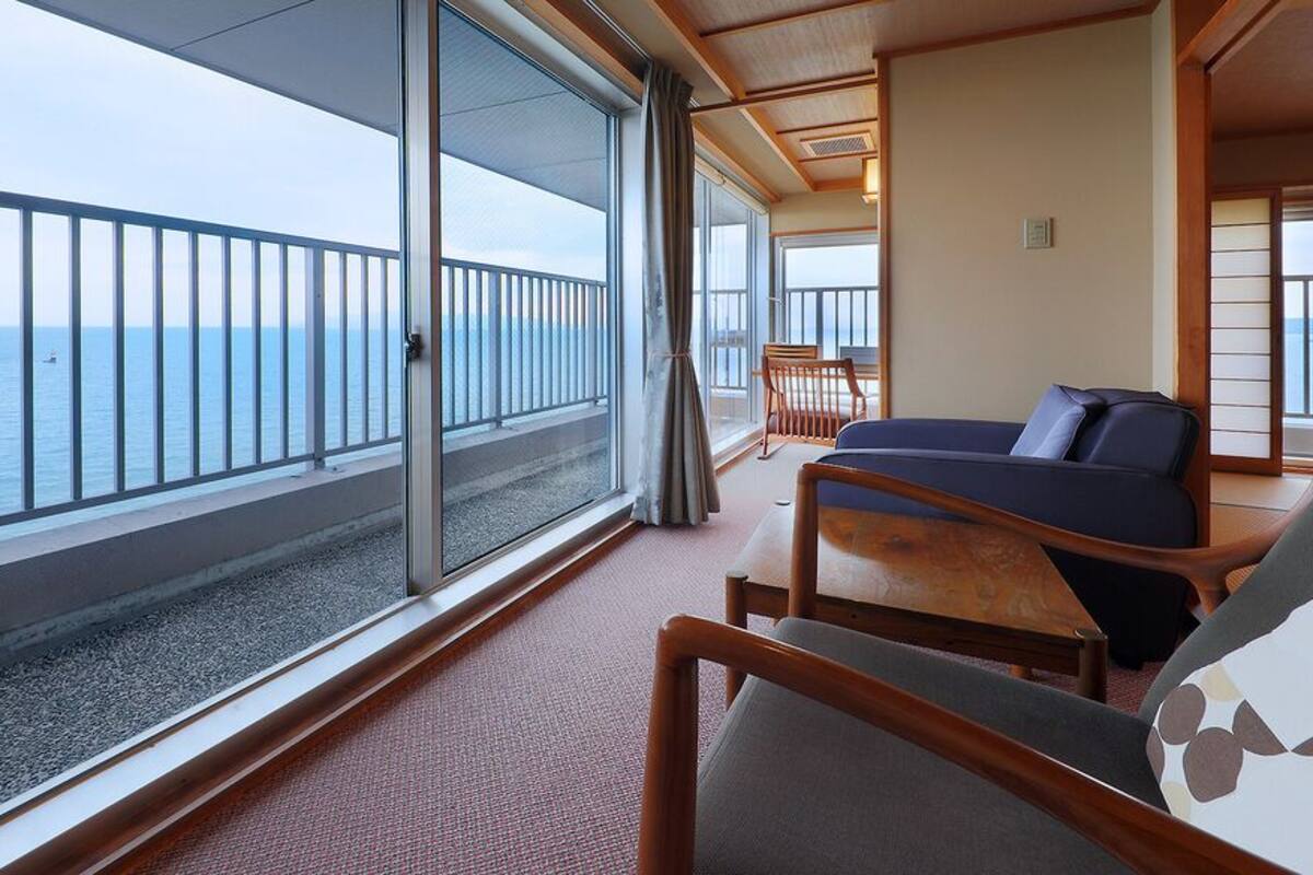 Een balkon met uitzicht op zee, een bed met een blauw hoofdeinde en een houten stoel.