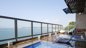 Piscina infinita con vistas al mar, terraza de madera, sillas de descanso y zona de estar cubierta.