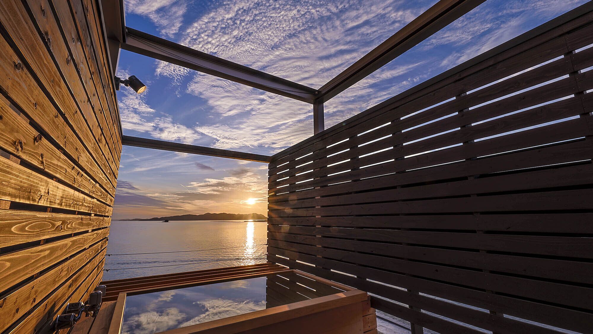 Een houten hutje met een glazen dak, een houten terras en uitzicht op de ondergaande zon over het water.