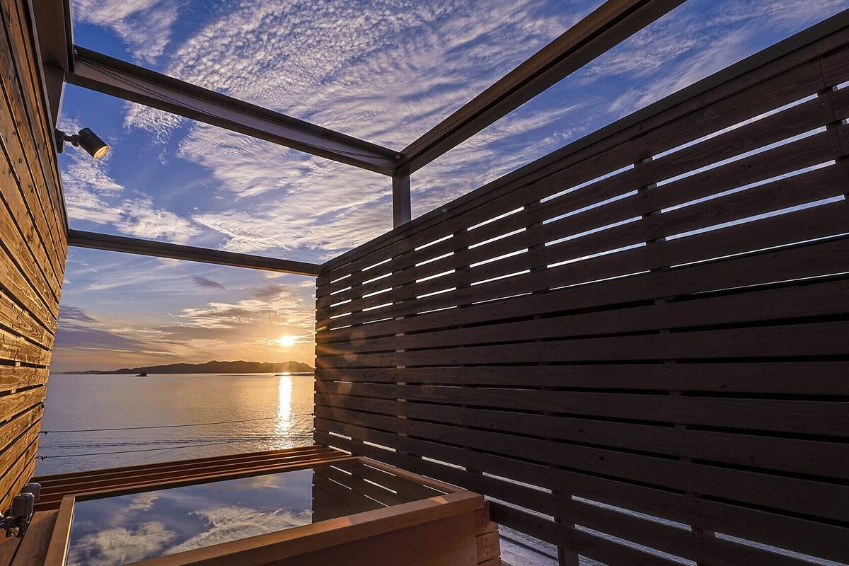 Een houten hutje met een glazen dak, een houten terras en uitzicht op de ondergaande zon over het water.