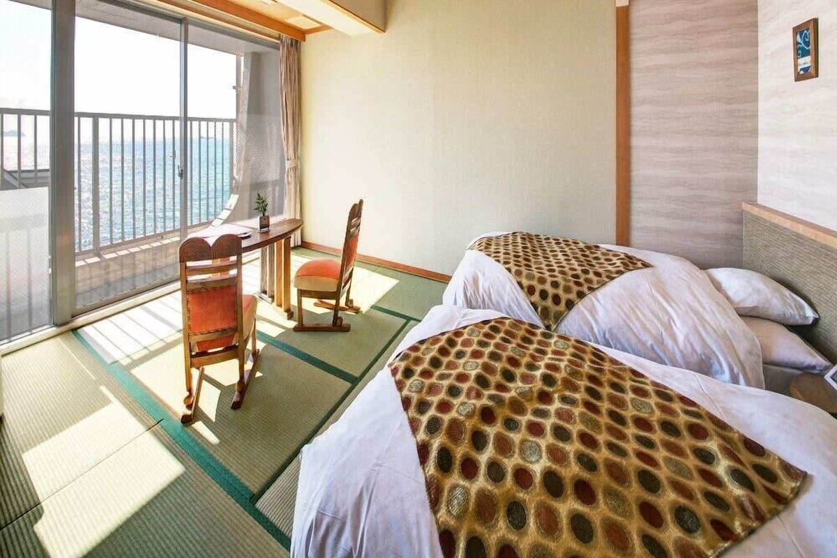 Een kamer met twee bedden, een bureau en een stoel, met uitzicht op zee door grote ramen.