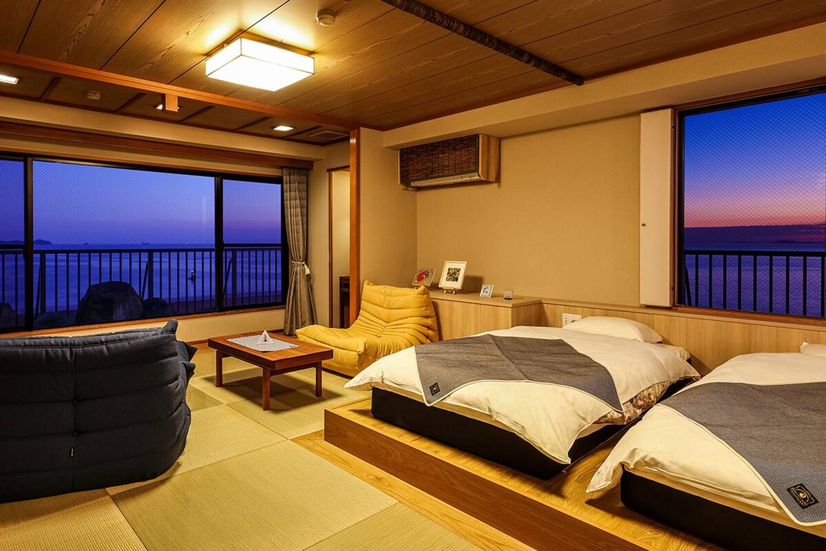 A room with two beds, a chair, a table, and a view of the sea.