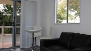 A modern room with a black leather sofa, a white table, and a sliding glass door leading to an outdoor area with greenery. - Huntsville Caravan Park (Tinana)