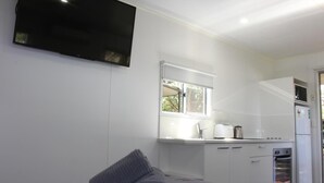 A modern, compact room with a bed, a flat-screen TV mounted on the wall, a window with a view, a kitchenette with a fridge, oven, and sink. - Huntsville Caravan Park (Tinana)