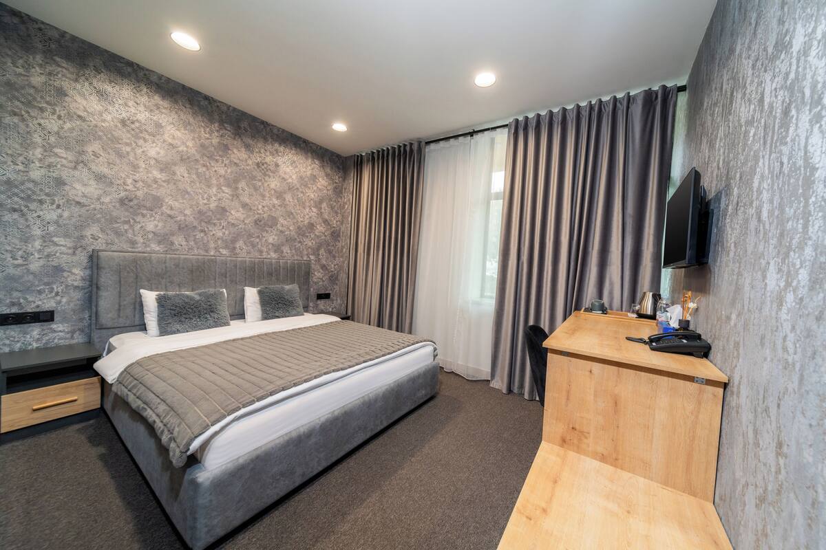 A modern hotel room with a large bed, a wooden desk, a flat-screen TV, and a large window with curtains.