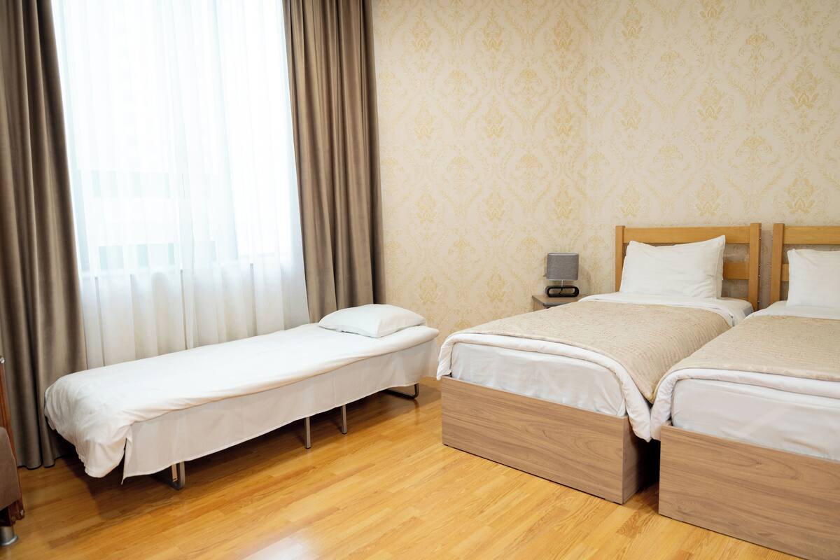 A hotel room with two single beds and one double bed, wooden flooring, a nightstand with a lamp, and a window with curtains.