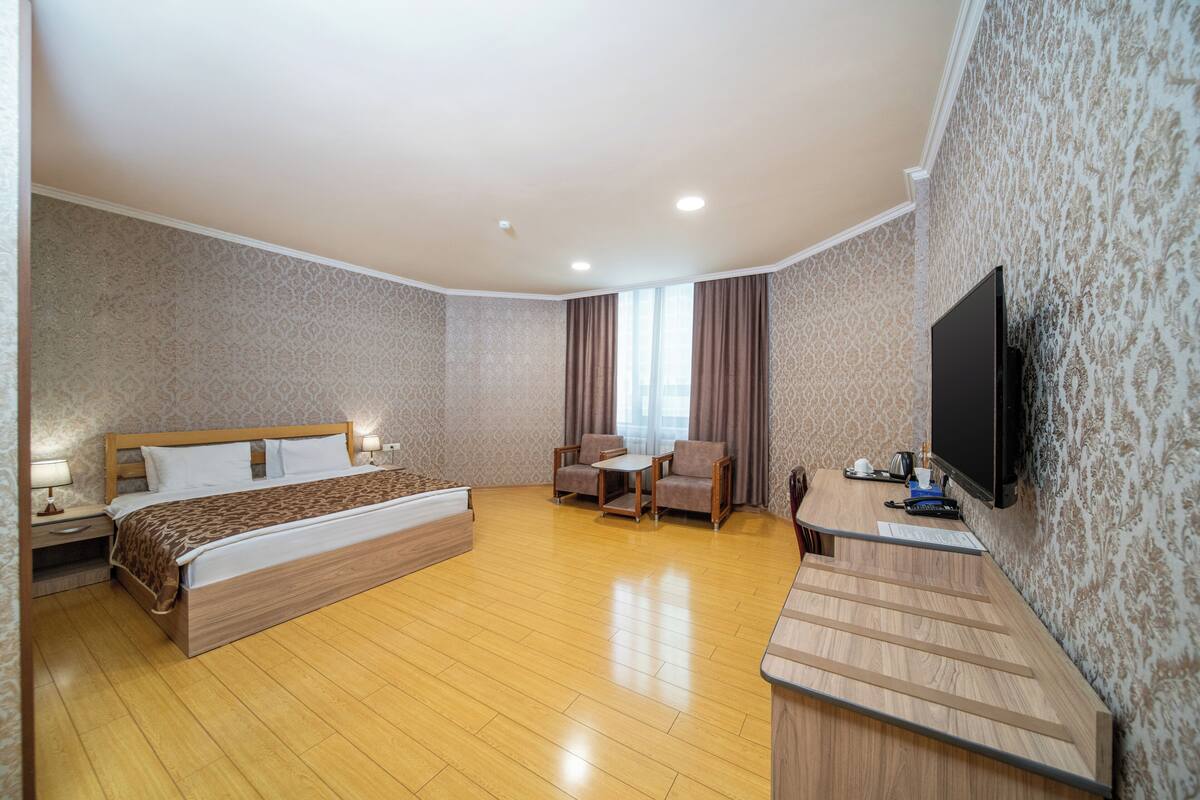 A hotel room with a large bed, a desk, two chairs, a TV, and a window with curtains.