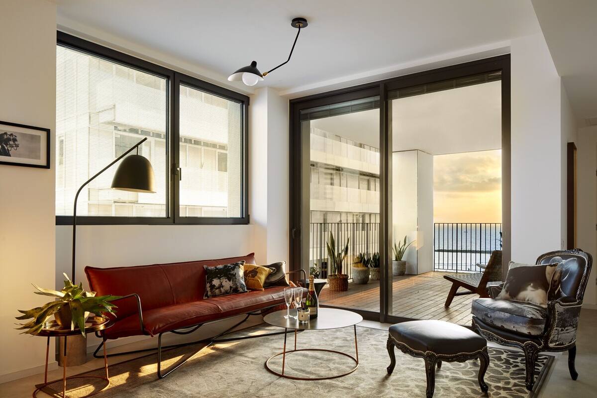 Una sala moderna con un sofá rojo, una mesa de centro redonda y un sillón de cuero. Hay ventanas amplias y vista al mar.