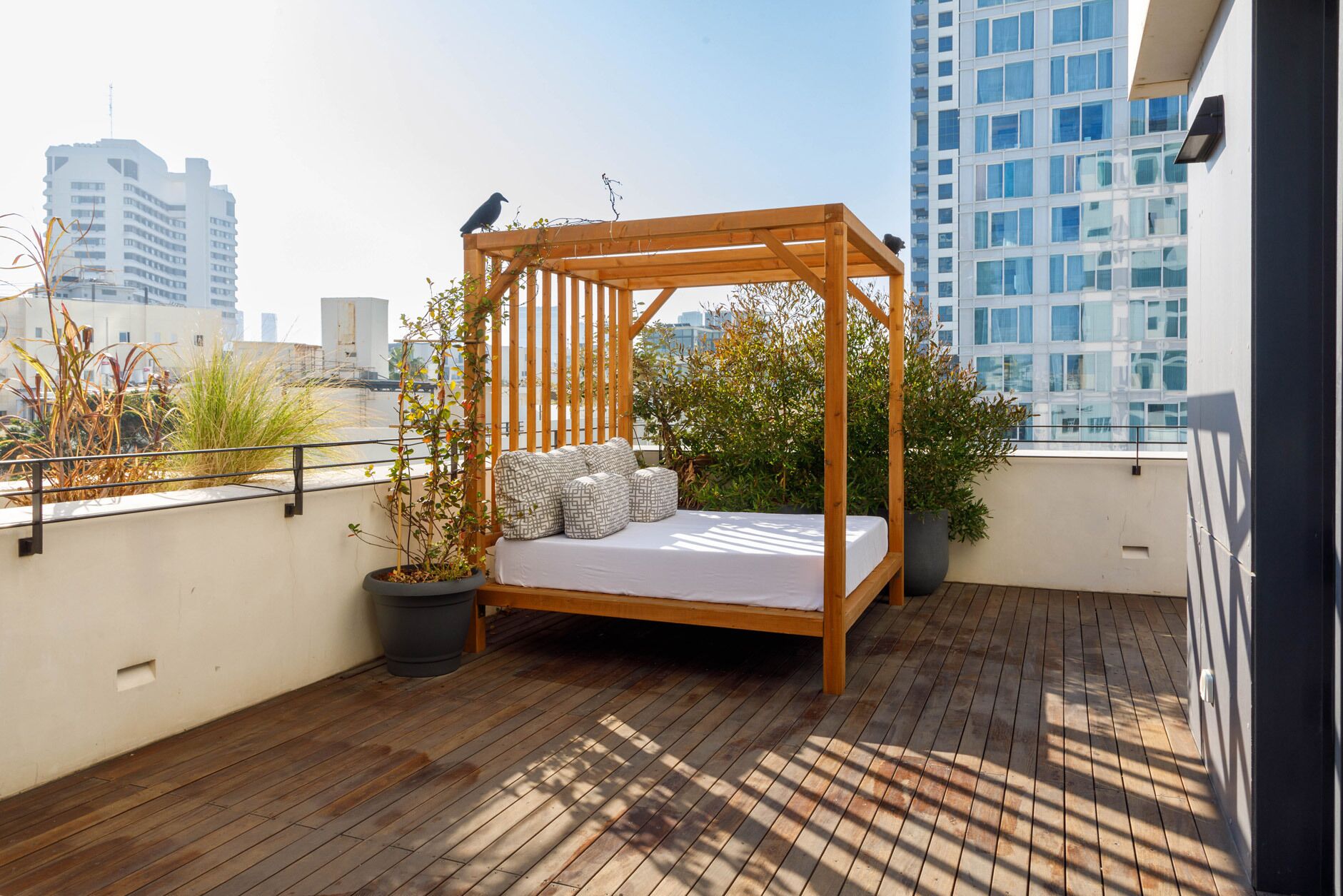 Una cama de madera con dosel en una terraza en la azotea, con plantas en macetas y vistas a edificios de la ciudad.