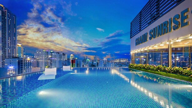 A modern hotel with a large infinity pool, high-rise buildings in the background, and a clear sky.