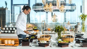 A chef preparing food in a modern kitchen with hanging pendant lights, buffet-style food stations, and a view of the city through large windows.