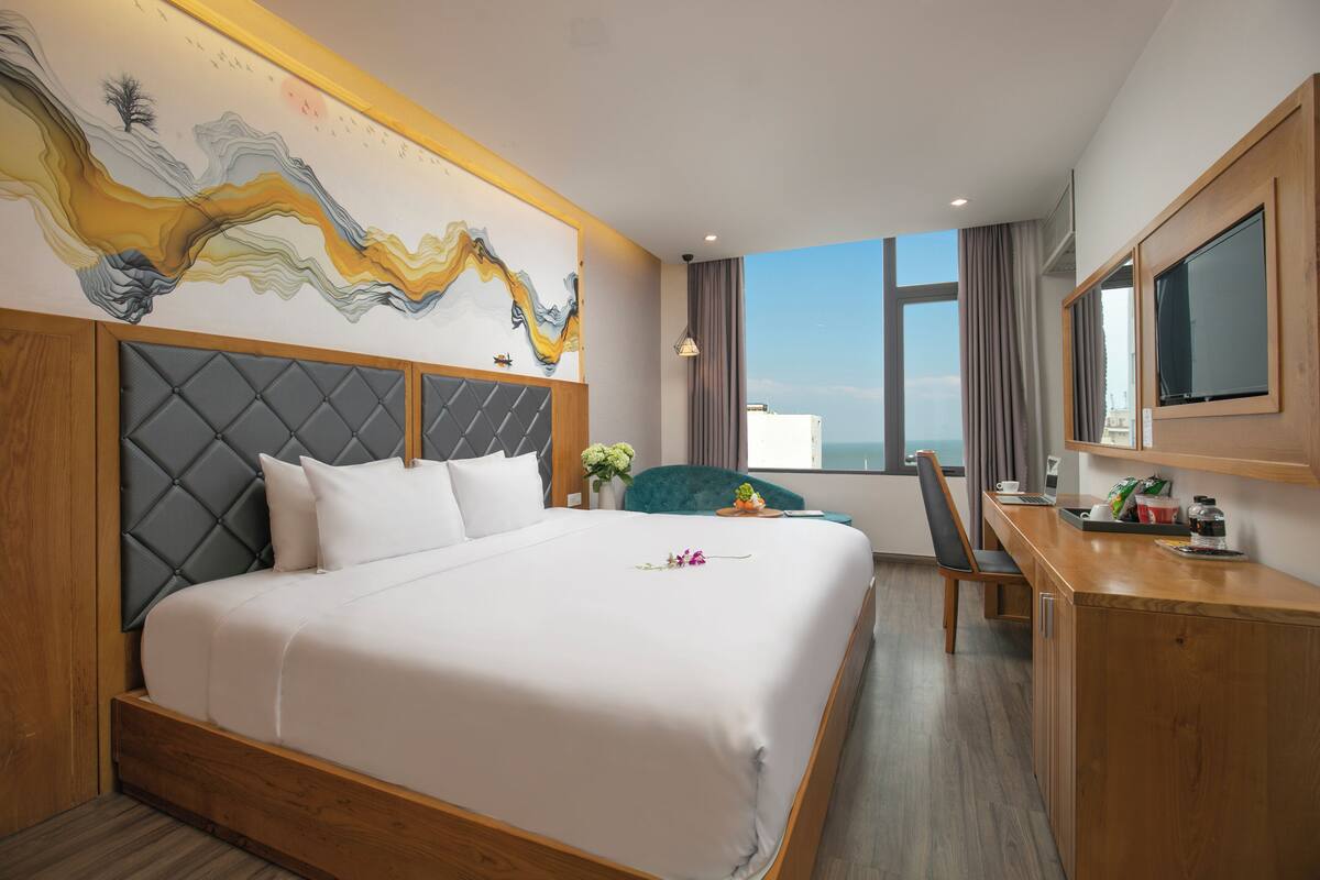 A hotel room with a large bed, a desk, a chair, a TV, and a window with a view.