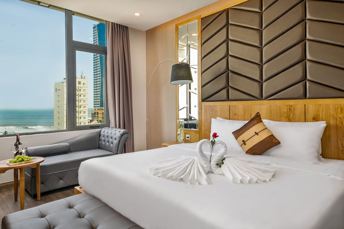 A hotel room with a large bed, a sofa, a small table with a bottle and glasses, and a view of the ocean.