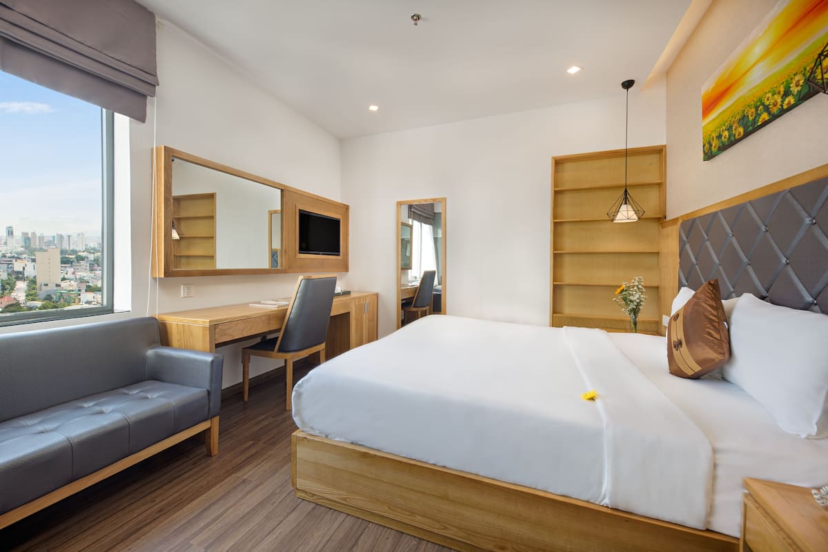 A modern hotel room with a large bed, a desk, a chair, and a sofa. There is a city view through the window, and a painting on the wall.