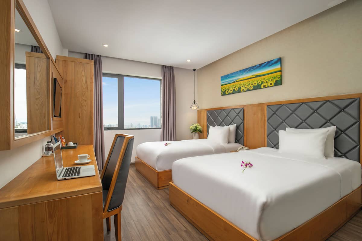 A hotel room with two beds, a desk with a laptop, a chair, and a large window offering a city view.