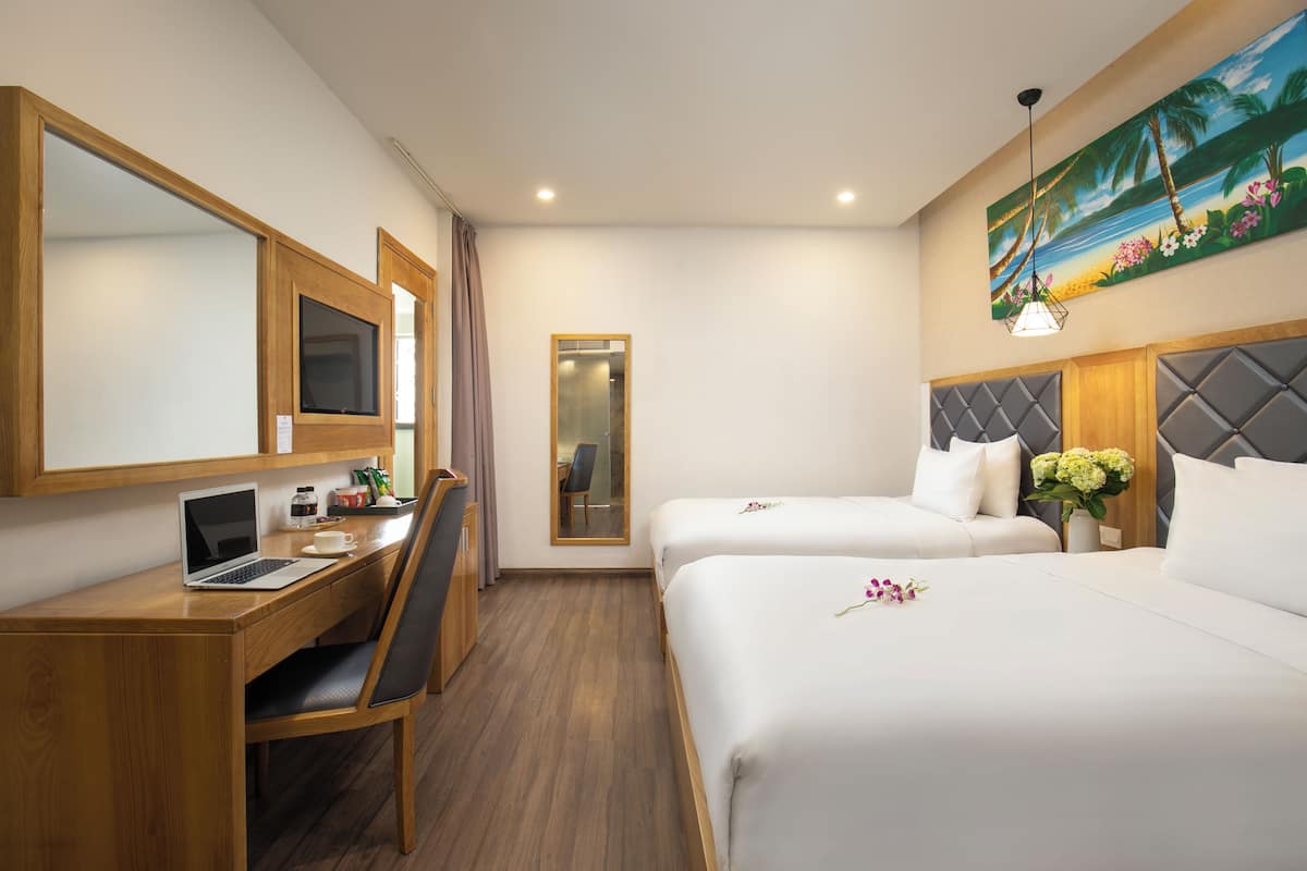 A hotel room with a wooden desk, a laptop, a chair, two beds, a mirror, a painting, and a small plant.