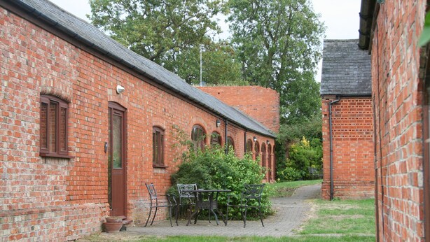 A brick building with a paved courtyard, outdoor seating, and a small tree. - Coldham Cottages (Bedford)