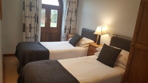 A hotel room with two beds, a wooden headboard, a nightstand with a lamp, and a window with curtains. - Coldham Cottages (Bedford)