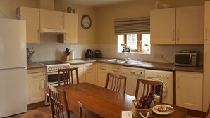 A compact kitchen with white cabinets, a wooden dining table, and basic appliances including a microwave, stove, and dishwasher. - Coldham Cottages (Bedford)