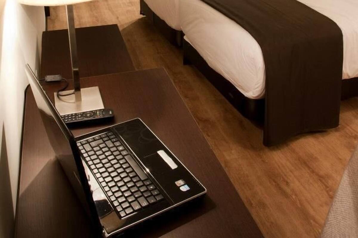 Quarto de hotel com cama, escrivaninha, laptop, luminária e guarda-roupa.
