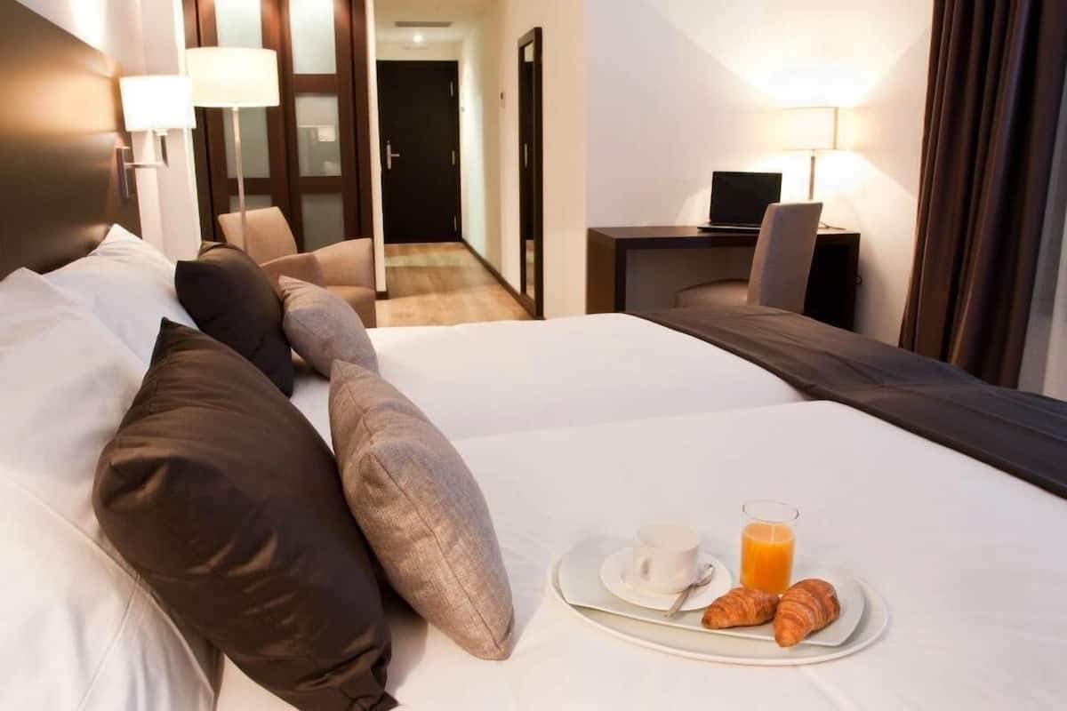 Quarto de hotel com cama, mesa de cabeceira com abajur, televisão e bandeja com croissants e suco.