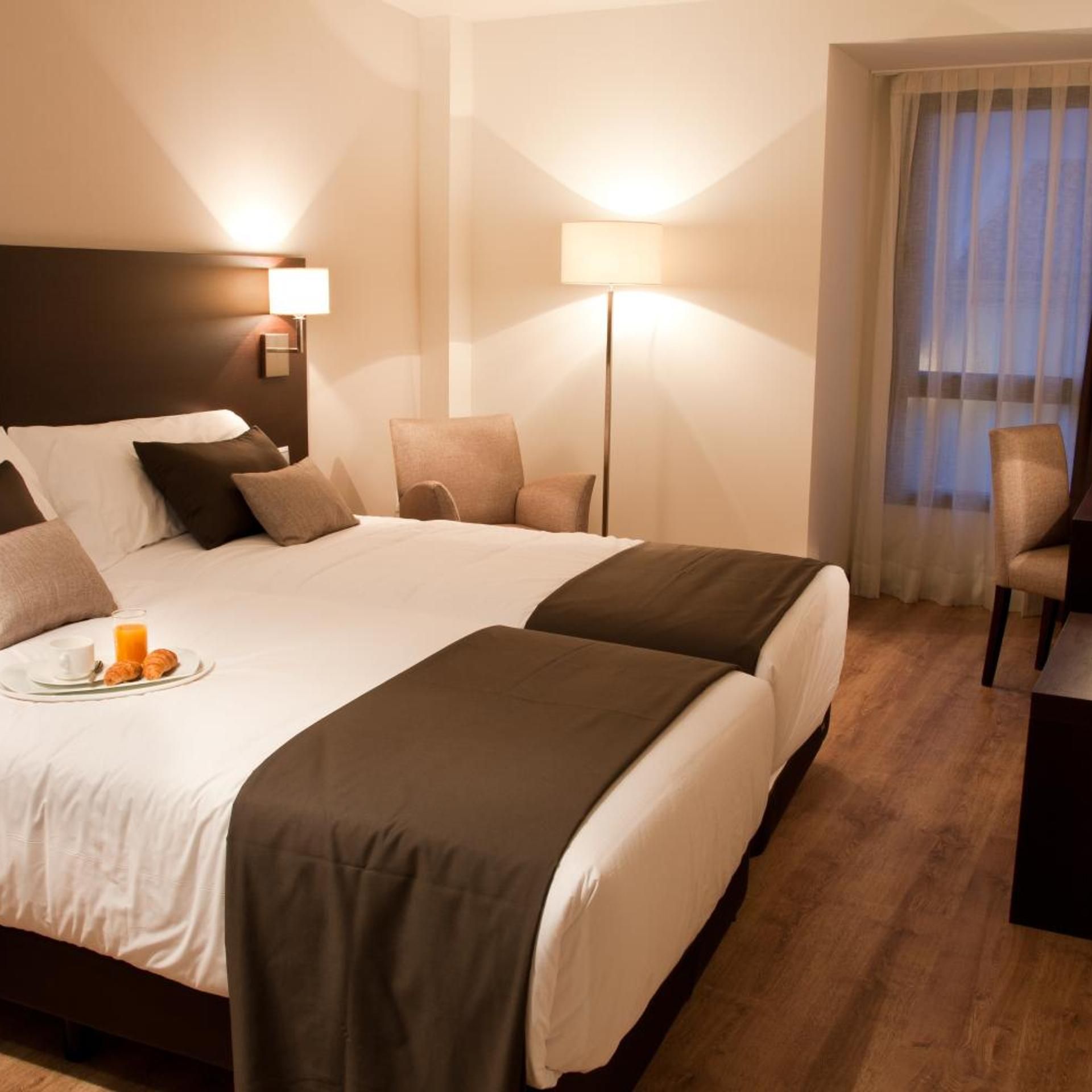 Habitació d'hotel amb un llit gran, dues taules de nit, una cadira, un escriptori i una finestra amb cortines.
