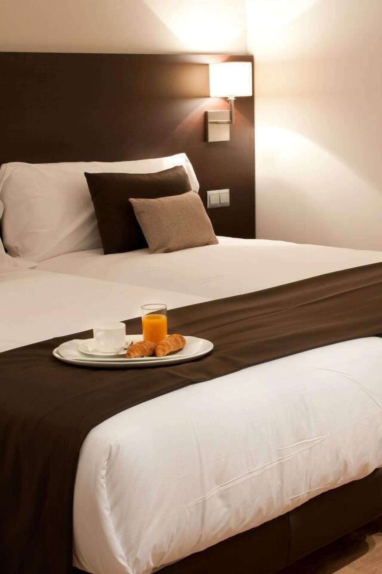 Une chambre d’hôtel avec un lit parfaitement fait, une table de chevet sur laquelle se trouvent un verre de jus d’orange et des croissants, et une lampe fixée au mur.