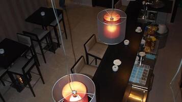 Un área de comedor con varias mesas y sillas, una barra con plancha, y lámparas colgantes.