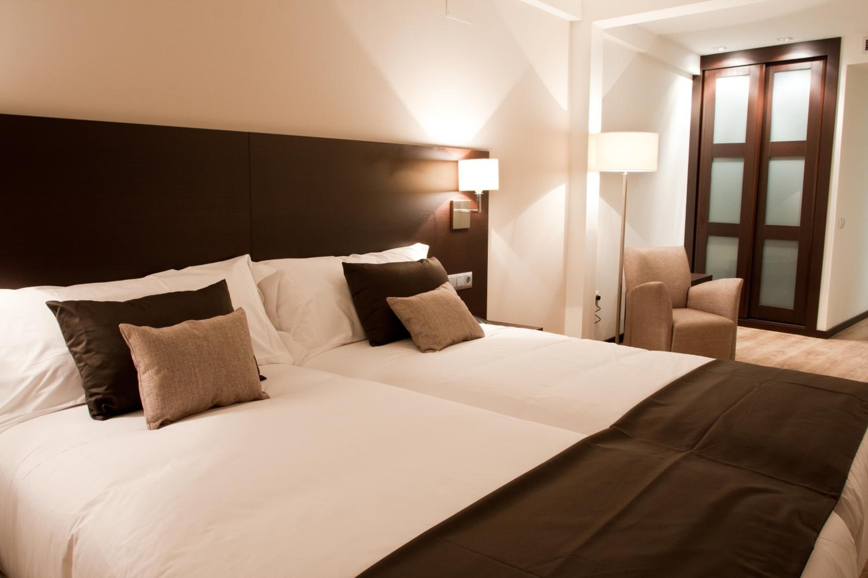 A hotel room with two beds, a dark headboard, a nightstand with a lamp, a chair, and a wardrobe with frosted glass doors.