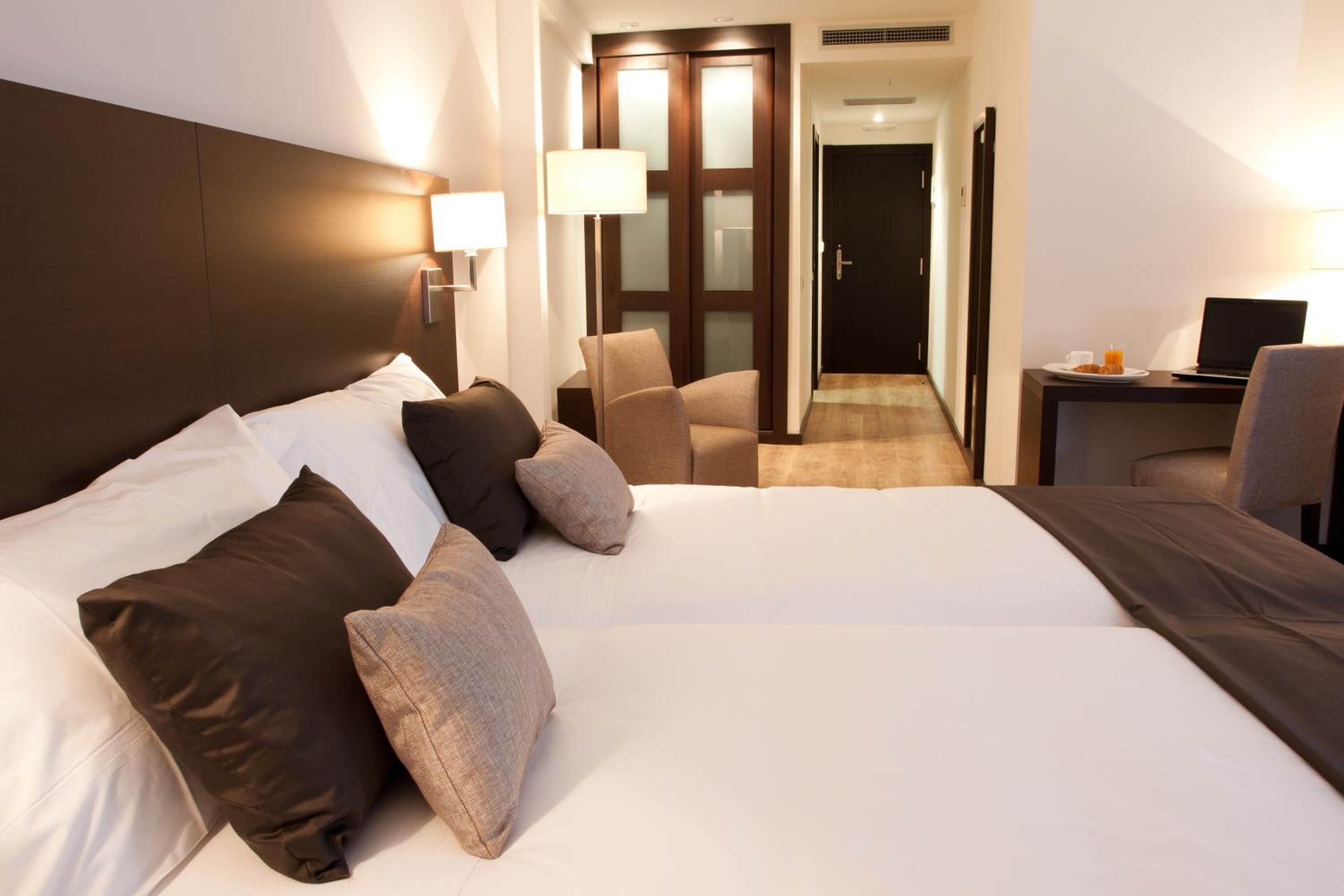 Habitación de hotel con una cama grande, un escritorio con una computadora portátil, una silla y una puerta que da a un pasillo.