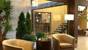 A lobby with two armchairs, a small table, and a potted plant. There is a staircase and a bar area visible through the glass wall. - Hotel Paloma (Tomelloso)