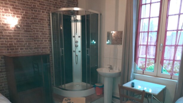 A bathroom with a glass-enclosed shower, a sink, a window with curtains, a wooden chair, and a small table with a remote control. - Auberge Des Pilotis (Beauvoir-en-Lyons)