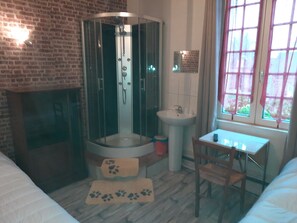 A bathroom with a glass-enclosed shower, a sink, a window with curtains, a wooden chair, and a small table with a remote control.