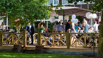 A covered outdoor seating area with wooden railings and tables, surrounded by trees and a building in the background.